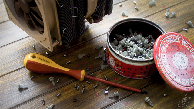 A wooden planked floor with a lot of items laying on it. In the top-left corner there's a large aluminium radiator with double brown and tan fans. At the bottom lies a red and orange Phillips screwdriver. On the right side is a short, but fat, opened red coffee can labeled "Uncle Gonzales' Whole Bean Coffee". Between the items there are a lot of nuts, bolts, screws and washers scattered around.
